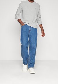 Harmaa pitkähihainen huppari, jossa on pyöreä kaula-aukko ja pieni logo, yhdistettynä löysiin sinisiin farkkuihin ja valkoisiin tennareihin.