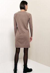 Robe en tricot marron clair, manches longues, coupe ajustée. Portée avec des collants noirs et des bottines. Texture lisse, col et poignets côtelés.