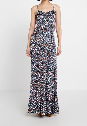 Mujer con vestido sin mangas hasta el tobillo, con estampado floral, tirantes finos y escote con volantes, de pie frente a un fondo blanco liso.