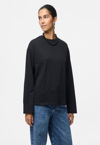 Μαύρη μπλούζα μακρυμάνικη από βαμβάκι, με στρογγυλή λαιμόκοψη και χαλαρή εφαρμογή. Φοριέται με μπλε τζιν παντελόνι και μαύρο κολιέ με χάντρες.