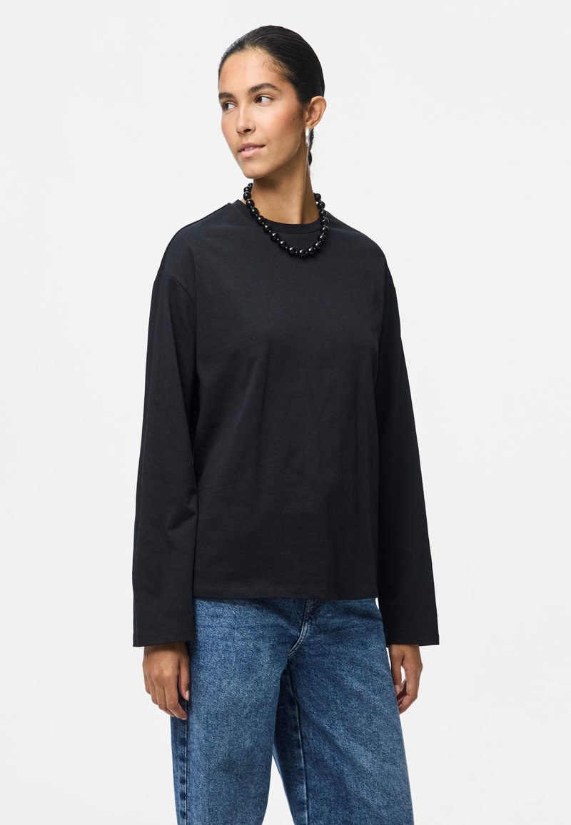 Μαύρη μπλούζα μακρυμάνικη από βαμβάκι, με στρογγυλή λαιμόκοψη και χαλαρή εφαρμογή. Φοριέται με μπλε τζιν παντελόνι και μαύρο κολιέ με χάντρες.