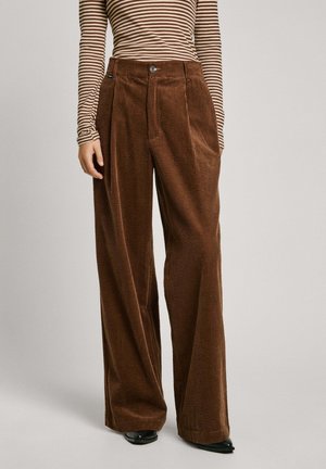 Kvinde iført højtaljede, vidde ben i brune kordfløjlsbukser kombineret med en brun og beige langærmet top med striber og sorte spidse sko.