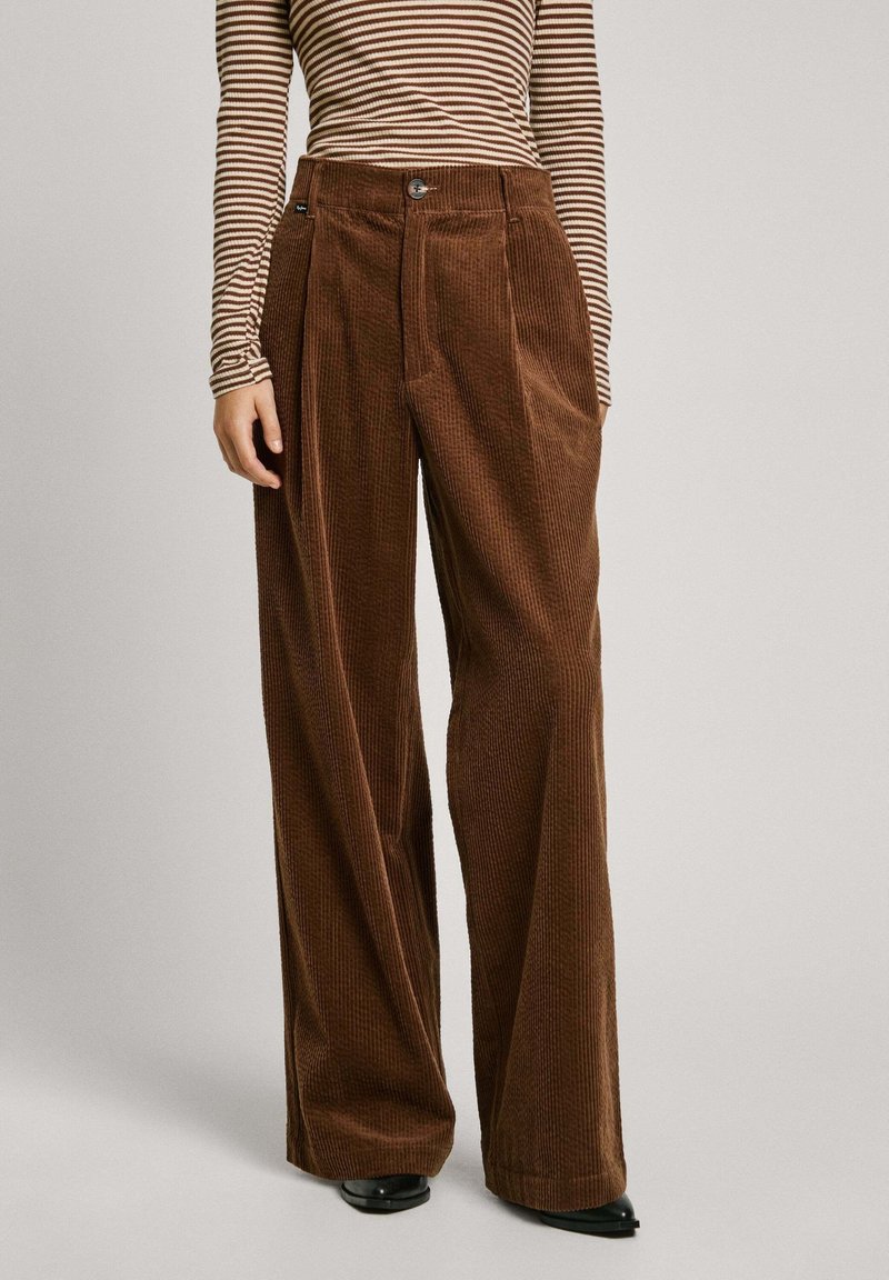 Femme portant un pantalon en velours côtelé marron taille haute à jambes larges, associé à un haut à manches longues rayé marron et beige, et des chaussures noires à bouts pointus.