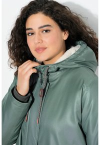 Grön vattentät jacka med en foderhuva i päls, utrustad med en flätad dragsko, brun accentknapp och texturerad tyg.