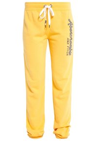 Žluté tepláky vyrobené z měkké tkaniny, s bílým šňůrkou na stažení, žebrovanými manžetami a šedým logem "Abercrombie AND FITCH" na boku.