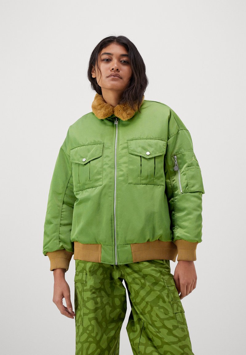 Giovane donna che indossa una giacca bomber verde con colletto in pelliccia marrone e pantaloni cargo mimetici verdi abbinati, in piedi davanti a uno sfondo neutro.