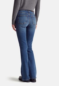 Jeans de corte acampanado en denim azul oscuro, con costuras visibles, dos bolsillos traseros y una cinturilla, combinados con zapatos negros.