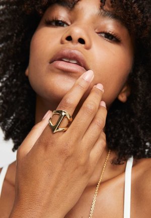 Anillo de oro con una forma única abierta y cortes angulares, usado en una mano con acabado de uñas naturales sobre piel clara.