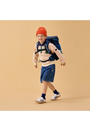 Young boy walking with a large backpack, wearing an orange beanie, striped long-sleeve shirt, denim shorts, and white sneakers.