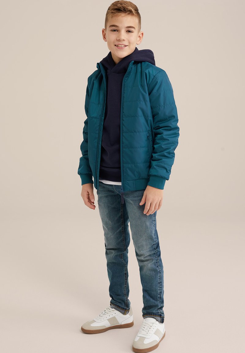 Teal vadderad jacka med en marinblå hoodie under, kombinerat med blå jeans och vita sneakers med ljusbruna detaljer. Stående position.