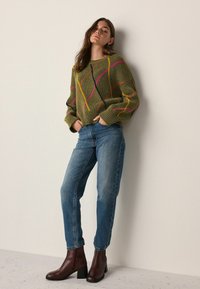 Green knit sweater with pink, yellow, and black diamond patterns, paired with medium-wash denim jeans and brown ankle boots.