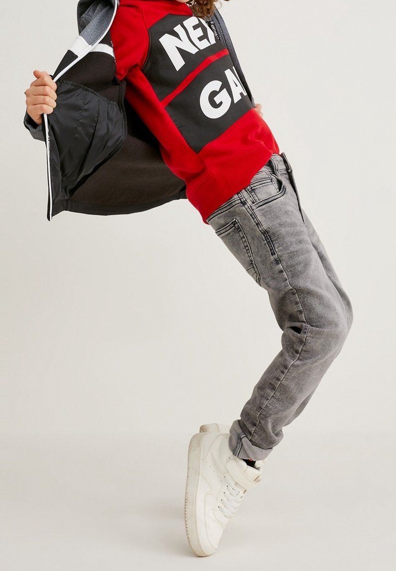 Enfant penchant en arrière portant un sweat-shirt rouge avec du texte, un jean gris, des baskets blanches, et tenant ouverte une veste sombre.