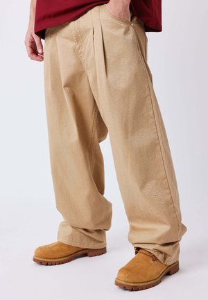 Personne portant un pantalon beige ample avec les mains dans les poches et des bottes en daim marron clair sur un fond uni clair.