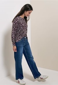 Frau in gemustertem Langarmshirt, mit weitem blauen Jeans und weißen Sneakers, die gegen einen geteilten beige und weißen Hintergrund vorwärts schreitet.