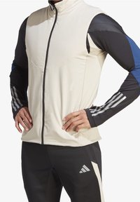 Chaleco atlético ajustado en beige con acentos en negro y azul. Cuenta con un cuello alto, cremallera completa y rayas en las mangas y los pantalones.