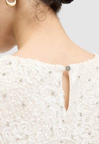 Hvid broderet kjole med detaljeret perlearbejde, en nøglehulsknaplukning bagpå og en struktureret stof med blomstermønstre.