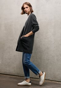 Grau, langer Cardigan mit Taschen, getragen über einem weißen Hemd. Dazu passen blaue, hochgekrempelte Jeans und weiße Sneakers mit schwarzen Akzenten.