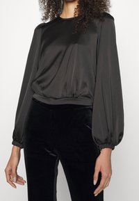 Donna che indossa una blusa di satin nera a maniche lunghe con maniche a sbuffo e pantaloni aderenti in velluto nero, in piedi contro uno sfondo bianco semplice.