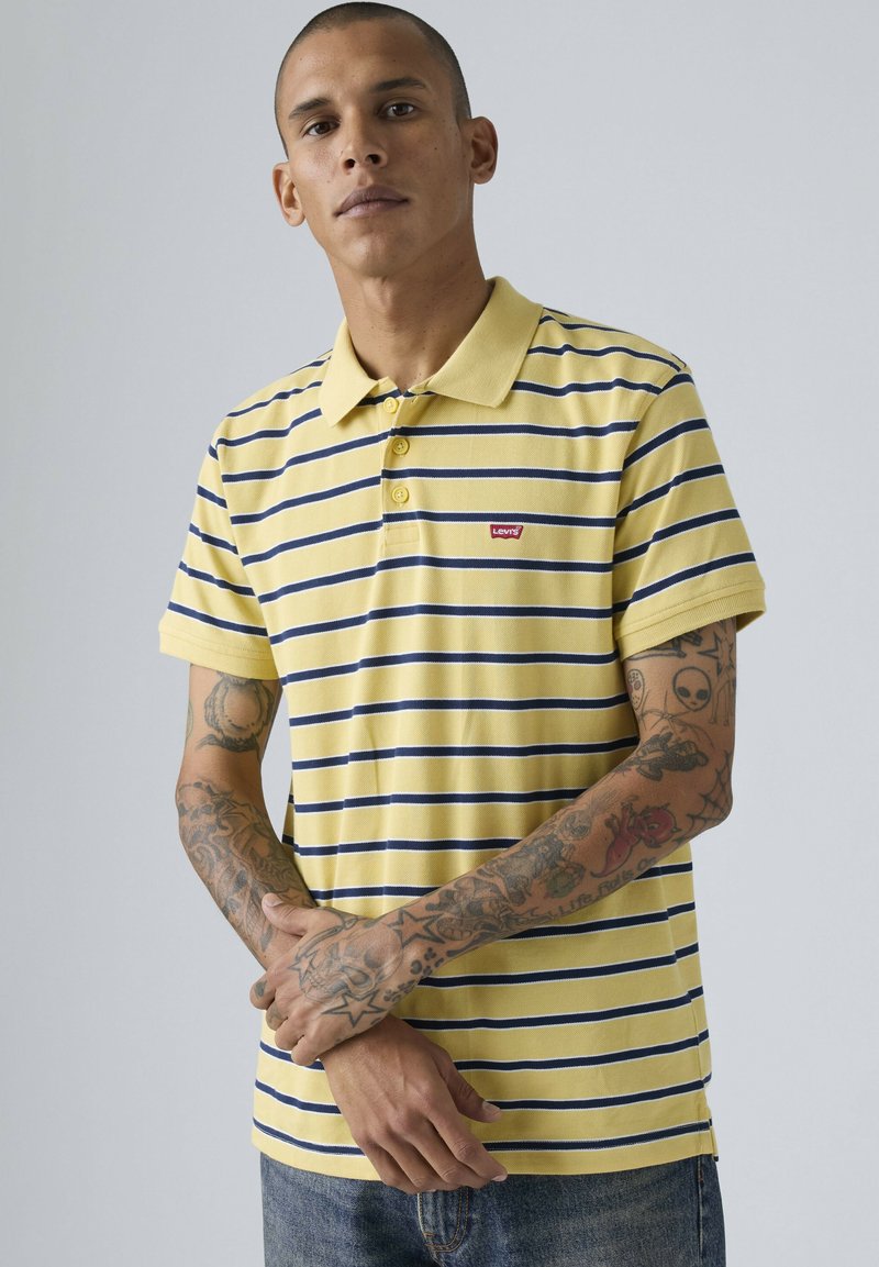 Homme aux bras tatoués portant un polo à manches courtes à rayures jaunes et bleu marine avec des boutons et un petit logo Levi's rouge sur la poitrine.