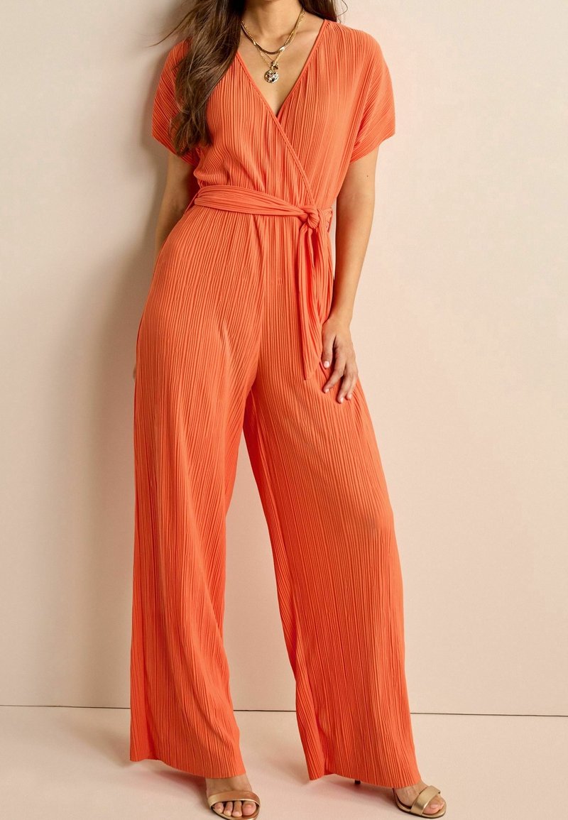 Femme portant une combinaison orange plissée à jambes larges et taille nouée, manches courtes, collier doré et sandales ouvertes, debout à l'intérieur.