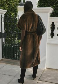 Brown faux fur coat with a belted waist, wide sleeves, and a soft texture, paired with black knee-high boots and a woven black handbag.