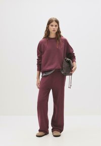 Felpa borgogna e pantaloni a gamba larga coordinati, realizzati in tessuto morbido. La modella tiene una borsa a tracolla grigia scura, in piedi su uno sfondo neutro.