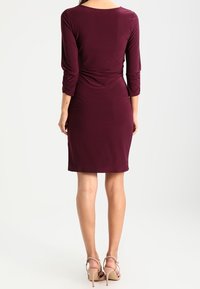 Viininpunainen, vartalonmyötäinen mekko, jossa on puolipitkät hihat, sivulla kerrostettu yksityiskohta ja polven pituinen helma, yhdistetty beigen värisiin korkeakorkoisiin sandaaleihin.