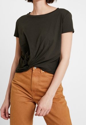 Kobieta ubrana w ciemnozielony krótki rękaw zawiązany w supeł, oraz jasnobrązowe spodnie z wysokim stanem, widoczna od szyi do bioder.