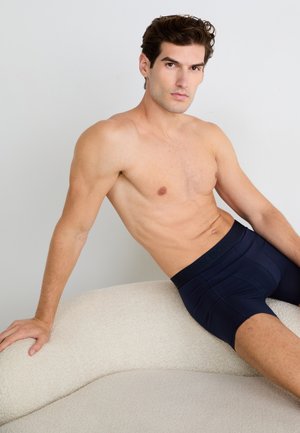 Ein shirtloser Mann mit kurzen dunklen Haaren sitzt auf einem strukturierten weißen, geschwungenen Sofa, trägt marineblaue Boxershorts und schaut in die Kamera.