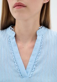 Ljusblå blus i strukturerad tygkvalitet med V-ringning och scalloped dekoration vid kragen. Mjuk, flödande material.