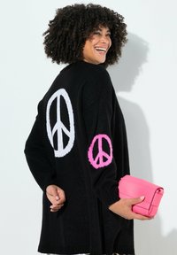 Sort strikket cardigan med store hvide og pink fredssymboler på ryggen og ærmerne. Modellen holder en lille pink taske.