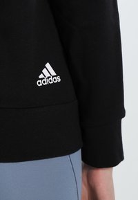 Sweat-shirt noir Adidas avec ourlet côtelé et logo blanc. Matière douce et extensible avec une coupe décontractée, associé à un legging bleu clair.