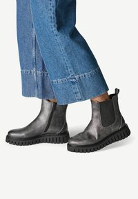 Des pieds chaussés de bottines en cuir gris métallisé avec des semelles noires épaisses et des panneaux latéraux en élastique, associées à un pantalon en denim bleu à jambes larges.