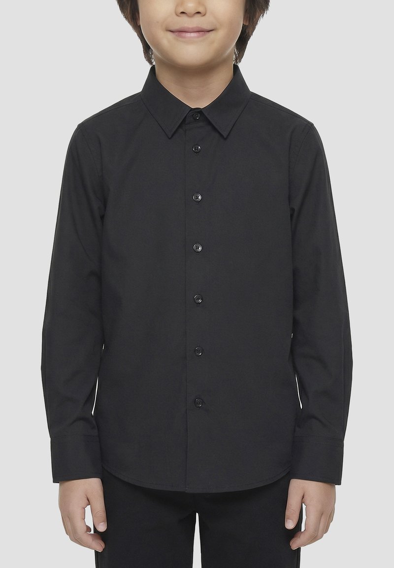 Enfant portant une chemise noire à manches longues boutonnée, debout avec les bras détendus devant un fond clair uni.