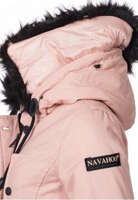 Lys rosa hættetrøje med faux fur kant, der har lynlåslukning, justerbare snore og et sort logo-patch på ærmet.