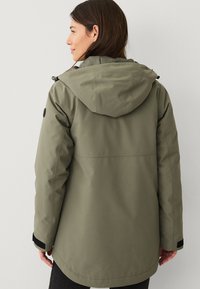 Donna che indossa una giacca a vento verde oliva con maniche regolabili, vista da dietro su uno sfondo bianco e semplice.