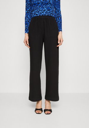 Pantalons larges noirs avec une taille élastique, texture lisse et détails de couture minimalistes, associés à des sandales à talons ouverts noires.