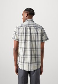 Camisa de manga corta a cuadros en crema claro con líneas gris oscuro, con cuello abotonado y dobladillo curvado. Tela de algodón texturizada.