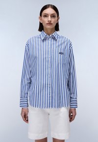 Camicia a righe blu e bianche con maniche lunghe, taschino sul petto e dettagli ricamati, abbinata a pantaloni corti bianchi. Tessuto dall'aspetto liscio.