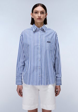 Camicia a righe blu e bianche con maniche lunghe, taschino sul petto e dettagli ricamati, abbinata a pantaloni corti bianchi. Tessuto dall'aspetto liscio.