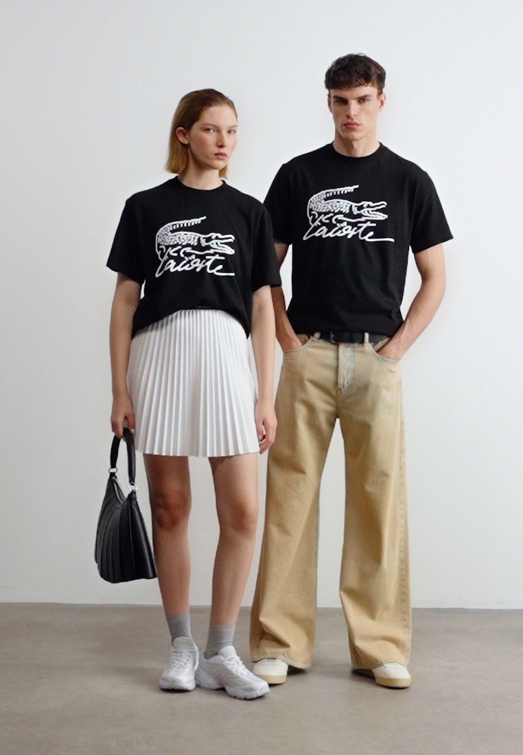 Жена и мъж, облечени в черни тениски с лого на Lacoste; жена в бяла плисирана пола и маратонки, държаща черна чанта; мъж в бежови панталони и маратонки.