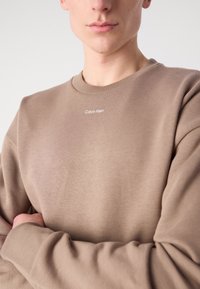 Béžová mikina s okrúhlym výstrihom vyrobená z mäkkého materiálu, s rebrovanými manžetami a lemom, s bielym logom "Calvin Klein" na hrudi.