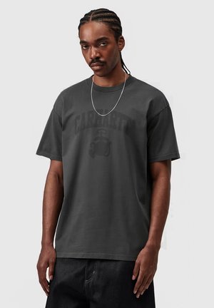 Uomo con capelli intrecciati che indossa una maglietta Carhartt grigio scuro e una collana di catena d'argento, in piedi davanti a uno sfondo chiaro e uniforme.