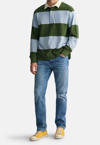 Groen en blauw gestreept poloshirt met lange mouwen en een crème-kleurige kraag, gecombineerd met lichtblauwe spijkerbroek en gele sneakers. Casual outfit.