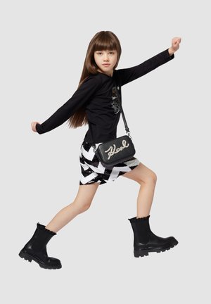 Haut noir à manches longues, jupe zippée noir et blanc à motifs zigzag, petit sac noir avec "Karl" en blanc, et bottines noires avec des bords volants.
