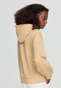 Felpa con cappuccio beige dal fit rilassato. Tessuto morbido e strutturato, polsini e orlo a coste. Presenta una tasca a marsupio e un design minimalista.