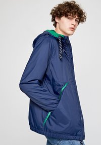 Giacca a vento blu navy con cappuccio verde, chiusura frontale con zip, polsini elastici e tasche laterali con dettagli verdi. Materiale liscio e leggero.