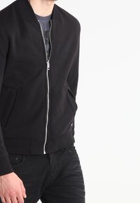 Svart zip-up jacka i slät tyg, med ståkrage, sidofickor och ribbade ärmslut. Minimalistisk design utan mönster.