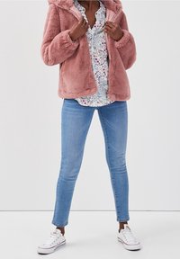 Person iført en lyserød, fuzzy jakke, blomstermønstret bluse, lyseblå slim fit jeans og hvide low-top sneakers, stående mod en hvid baggrund.