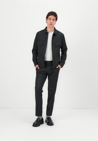 Schwarze Jacke mit Fronttaschen, getragen über einem weißen Hemd; kombiniert mit dunklen Hosen und schwarzen Schuhen, vor einem schlichten Hintergrund.